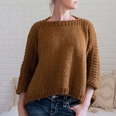 a woman standing in front of a white brick wall wearing a brown sweater and jeans