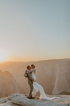 a bride and groom standing on top of a mountain at sunset with the sun setting behind them