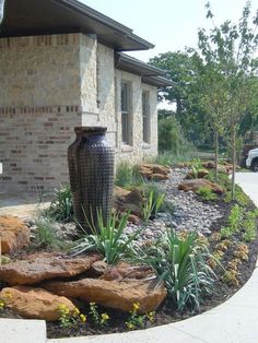 a large vase sitting in the middle of a flower bed next to a house with rocks and plants around it