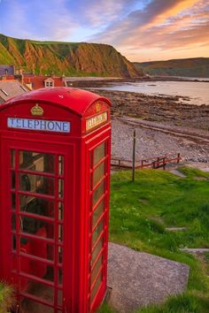 a red phone booth sitting on top of a lush green field next to the ocean