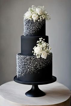 a three tiered black cake with white flowers on top and sprinkles