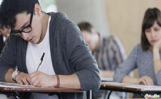 a man sitting at a desk writing in a classroom with two other people behind him