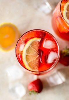 two glasses filled with fruit and ice on top of a white table next to strawberries