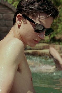 a young man in sunglasses sitting on the edge of a swimming pool while another person swims behind him