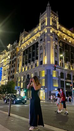 a woman is standing on the sidewalk in front of a large building at night time