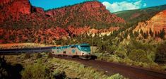 a blue and white train traveling through a lush green forest filled mountain side covered in red rocks