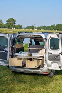 the back end of a silver van parked on top of a lush green field next to a lake