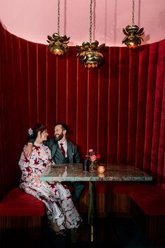 a man and woman sitting at a table in front of a red velvet wall with gold chandeliers