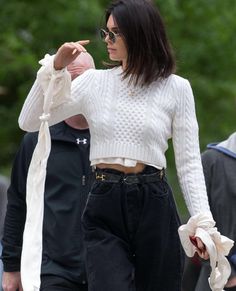 a woman in white sweater and black pants pointing at something on the ground with other people behind her