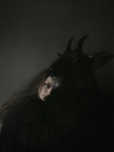 a woman with long hair wearing a demon mask and black coat, standing in the dark