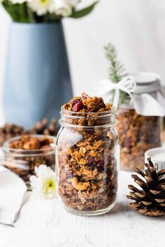 granola in a glass jar next to two small pine cones and a blue vase with white flowers