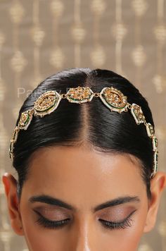 a woman wearing a gold and green headpiece with jewels on her forehead, looking down at the ground