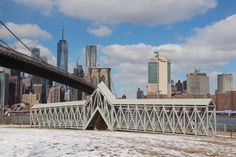 a bridge over the water in front of a cityscape with tall buildings and snow on the ground