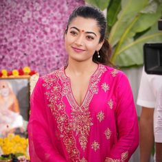 a woman standing in front of a flower arrangement wearing a pink outfit and gold jewelry