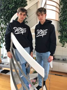 two young men standing next to each other in front of a spiral stair case with plants