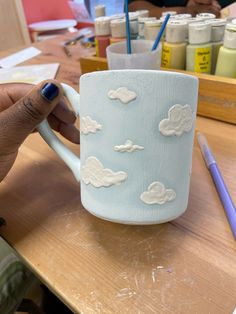 a person is holding a cup with clouds on it and paintbrushes in the background