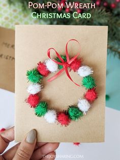 a hand holding a christmas card with pom - pom wreath on it