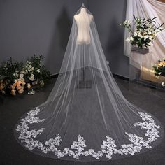 a white wedding veil with flowers on the bottom is hanging from a wall in front of a flower arrangement