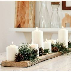 some white candles are sitting on a wooden tray with pine cones and greenery in front of them