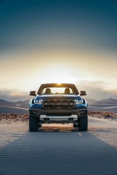 the front end of a blue pickup truck on a desert road at sunset with mountains in the background