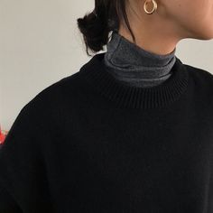 a woman wearing a black turtle neck sweater and large gold hoop earrings, standing in front of a white wall