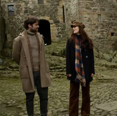 a man and woman standing next to each other on a cobblestone street in front of an old stone building