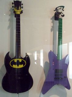 two guitars with batman logos on them are displayed in a glass case, one is purple and the other is black