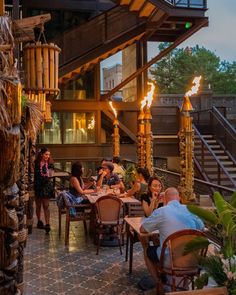 people are sitting at tables in an outdoor restaurant with fire torches on the outside wall