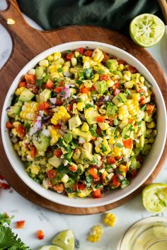 a white bowl filled with corn salad on top of a wooden cutting board next to lime wedges