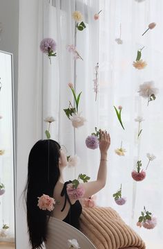 a woman sitting on top of a chair in front of a window with flowers hanging from it