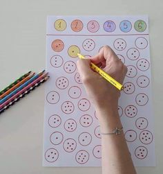 a person is holding a pencil and writing numbers on a sheet of paper with colored crayons next to it