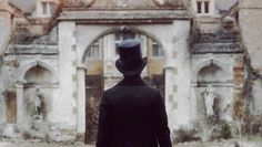 a man in a black suit and top hat walking towards an old building