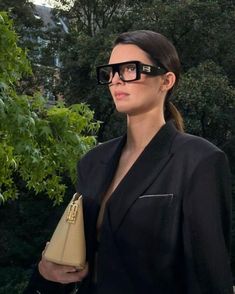 a woman in black suit and sunglasses holding a tan purse with trees in the background