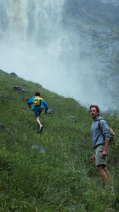 two men hiking up a hill in front of a waterfall with mist coming off the side