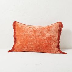 an orange velvet pillow with fringes on the front and back, against a white wall