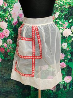 a mannequin wearing a white apron with red trim and flowers in the background