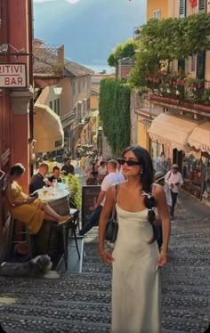 a woman in a white dress walking down a cobblestone street with people sitting at tables