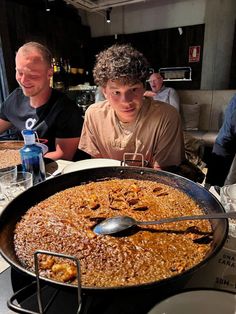 two men sitting at a table with a large pan of food in front of them