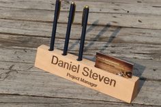 a wooden business card holder with four black pens in it on a wood table top