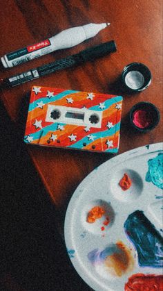 a wooden table topped with lots of different types of paints and brushes next to a paper plate