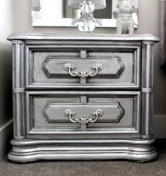 a silver dresser sitting next to a mirror on top of a table with a lamp