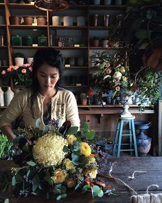 a woman arranging flowers in a vase on top of a wooden table next to shelves filled with potted plants