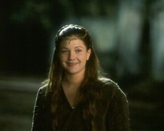 a woman with long hair wearing a brown sweater and smiling at the camera while standing in front of a dark background