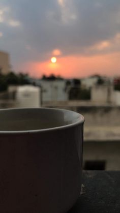 a white cup sitting on top of a wooden table next to a window with the sun setting in the background