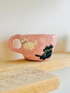 a pink coffee cup with cats on it sitting on top of a wooden shelf next to a white wall