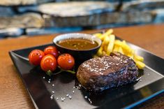 a steak and french fries on a black plate with tomatoes, sauce and ketchup