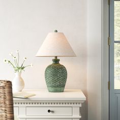 a green vase sitting on top of a white table next to a lamp and wicker basket