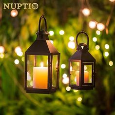 two hanging lanterns with lit candles in front of some trees and lights on the ground