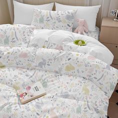 an unmade bed with white sheets and colorful floral designs on it, next to a pink teddy bear