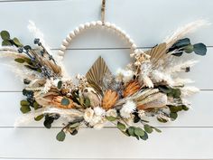 a wreath with dried flowers and leaves hanging on a white wooden wall next to a bead necklace